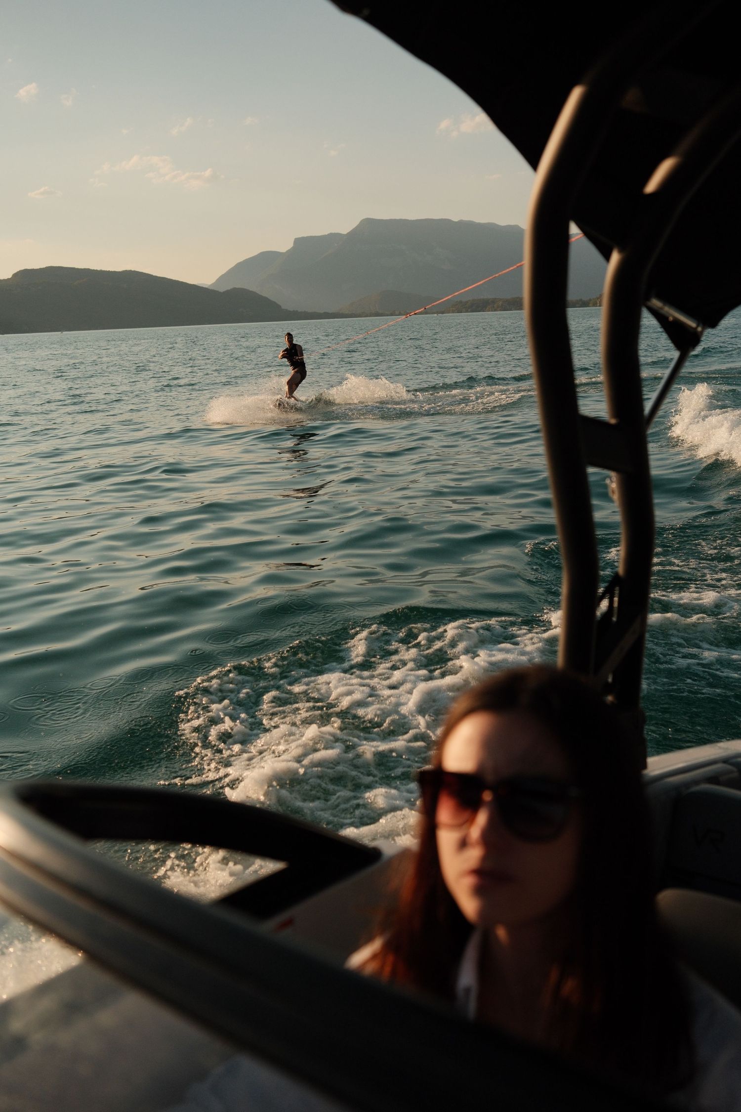 Woman in sunglasses on a speedboat watches a wakeboarder ride waves on a scenic mountain lake at sunset.