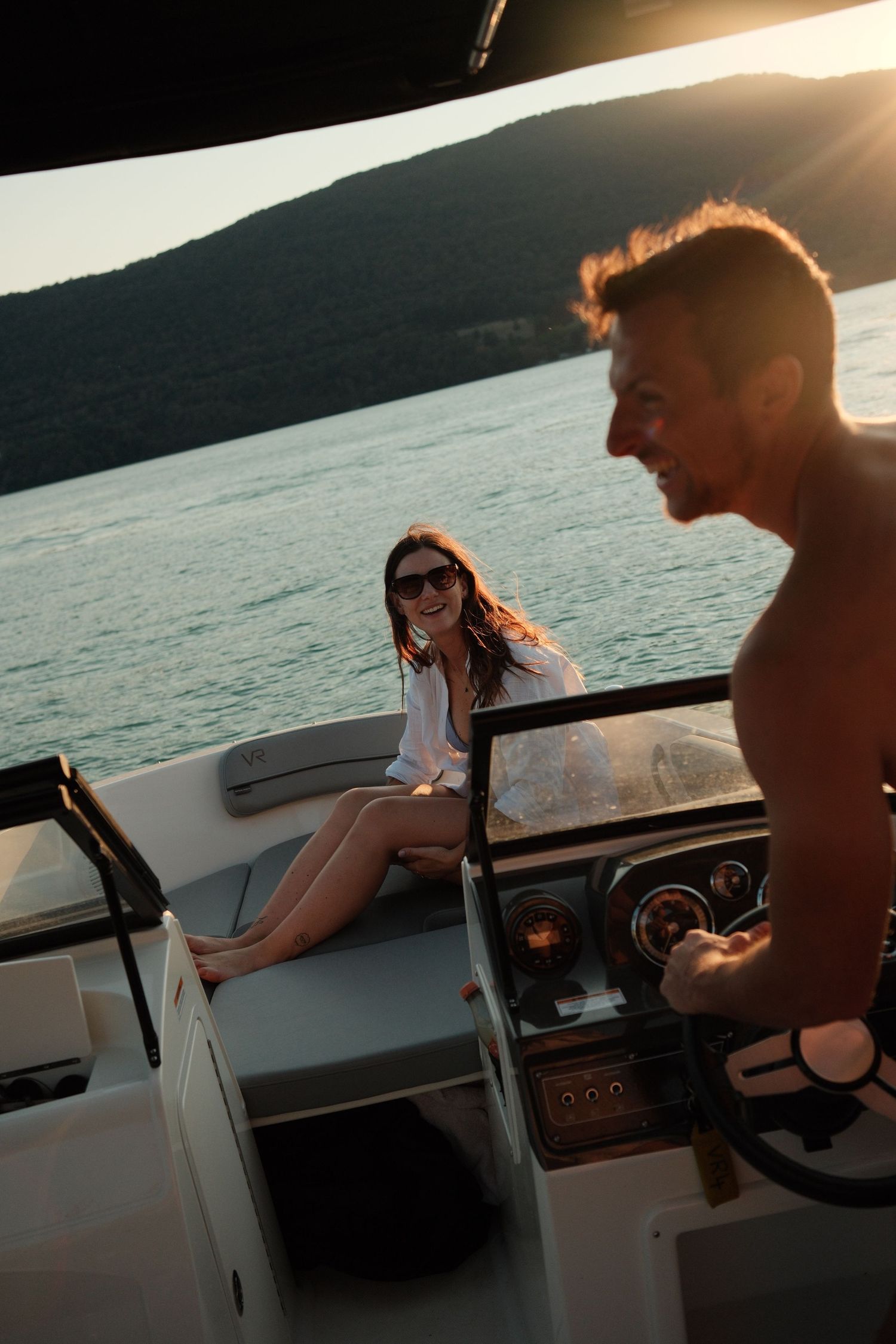 A smiling woman in sunglasses sits on a yacht deck while a shirtless man steers at sunset on calm waters.