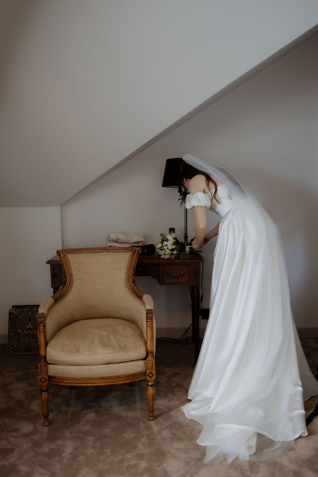 Bride in white off-shoulder gown with veil arranging flowers on antique desk beside vintage armchair.