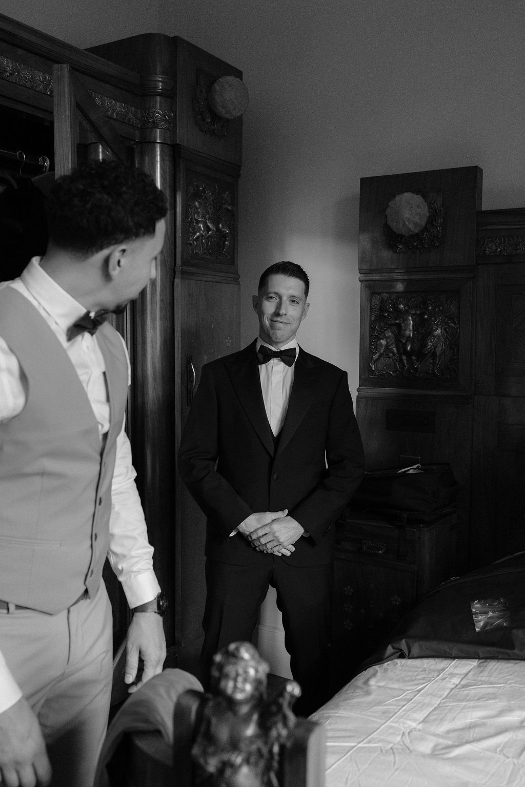Groom in black tuxedo with bow tie standing in ornate room with groomsman, black and white.