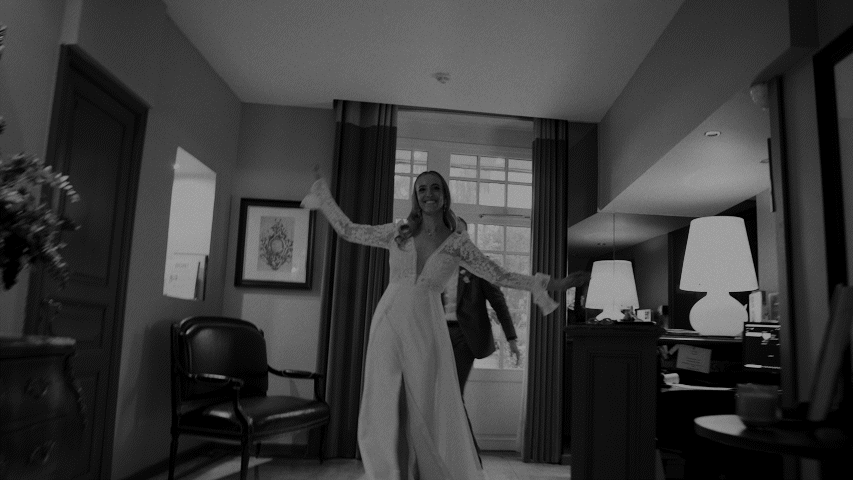 A woman in a flowing white gown poses dramatically in a dimly lit room in black and white photography.