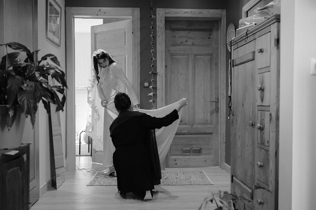 Black and white photo of a person kneeling to adjust a bride's wedding gown train in an elegant hallway.