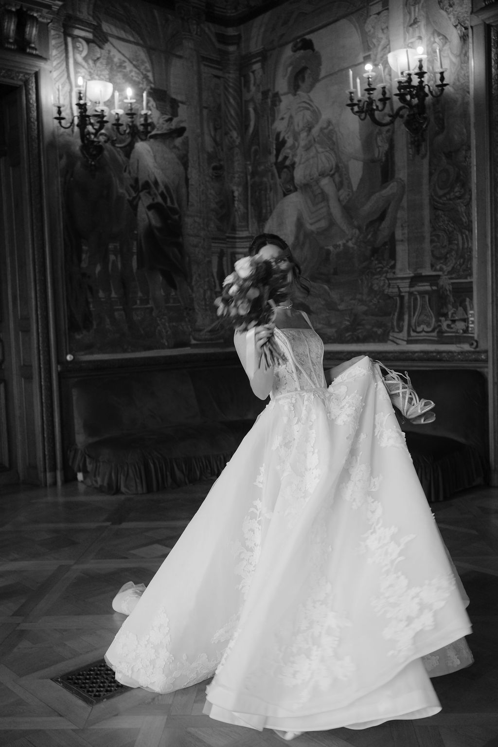 Black and white photo of bride twirling in lace ball gown holding bouquet in grand ornate ballroom.