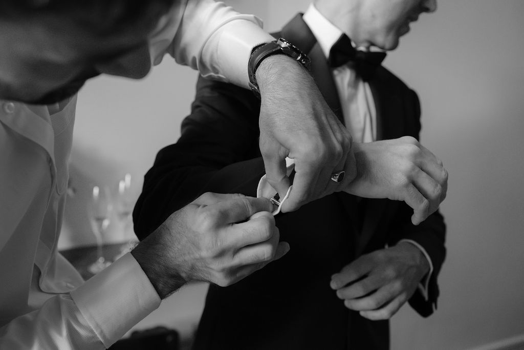 Groom getting cufflinks fastened by helper in black tuxedo at wedding preparation.