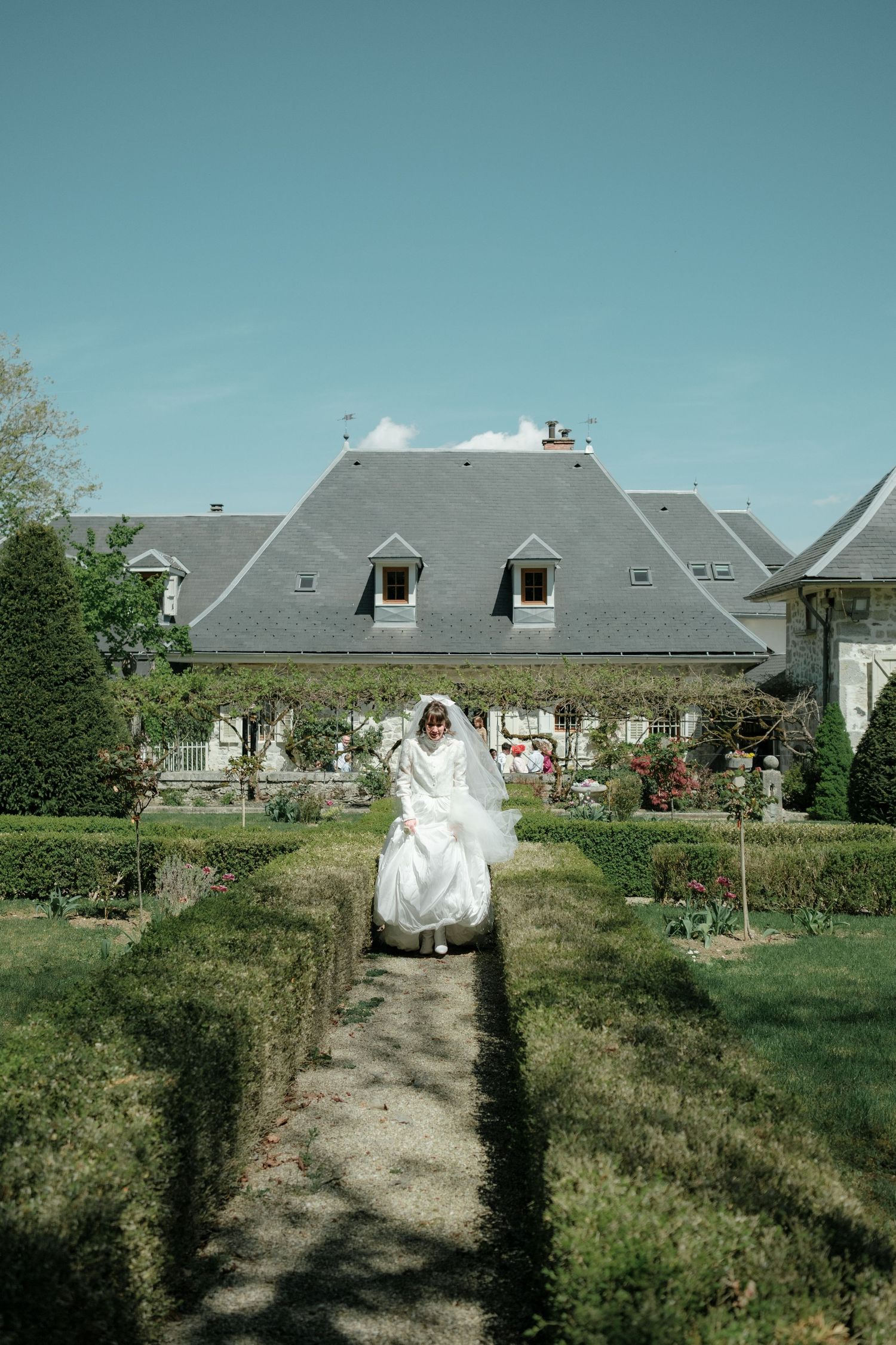 Bride in white gown and veil walks through formal garden hedges toward elegant French manor house on sunny day.