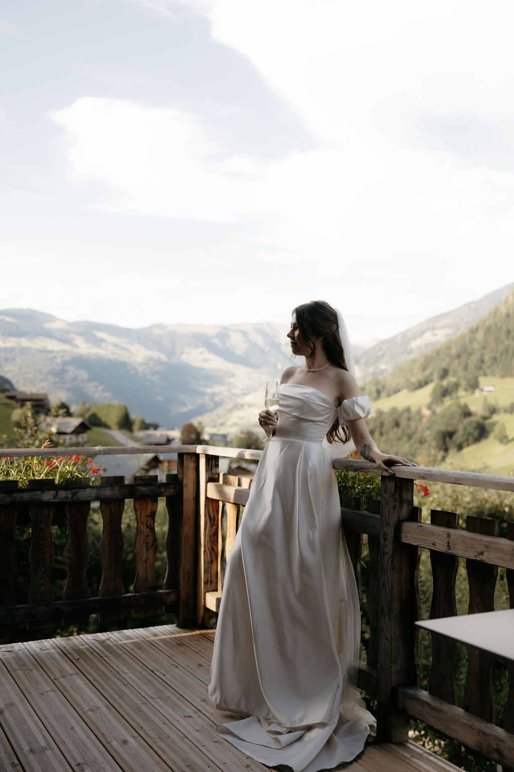 Bride in elegant white off-shoulder gown holding champagne on wooden balcony with mountain views.