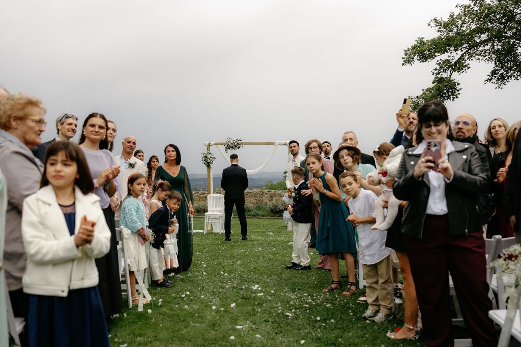 Wedding ceremony guests lining aisle outdoors on green lawn with floral arch backdrop.