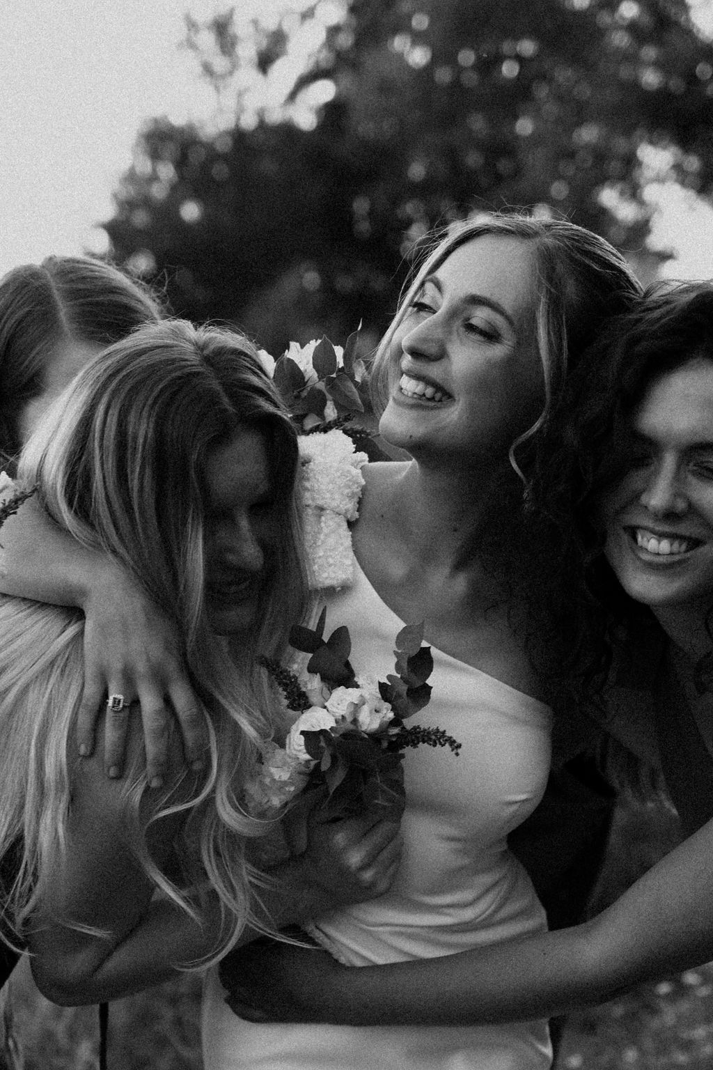 Black and white photo of smiling bride embraced by bridesmaids holding floral bouquet at outdoor wedding.