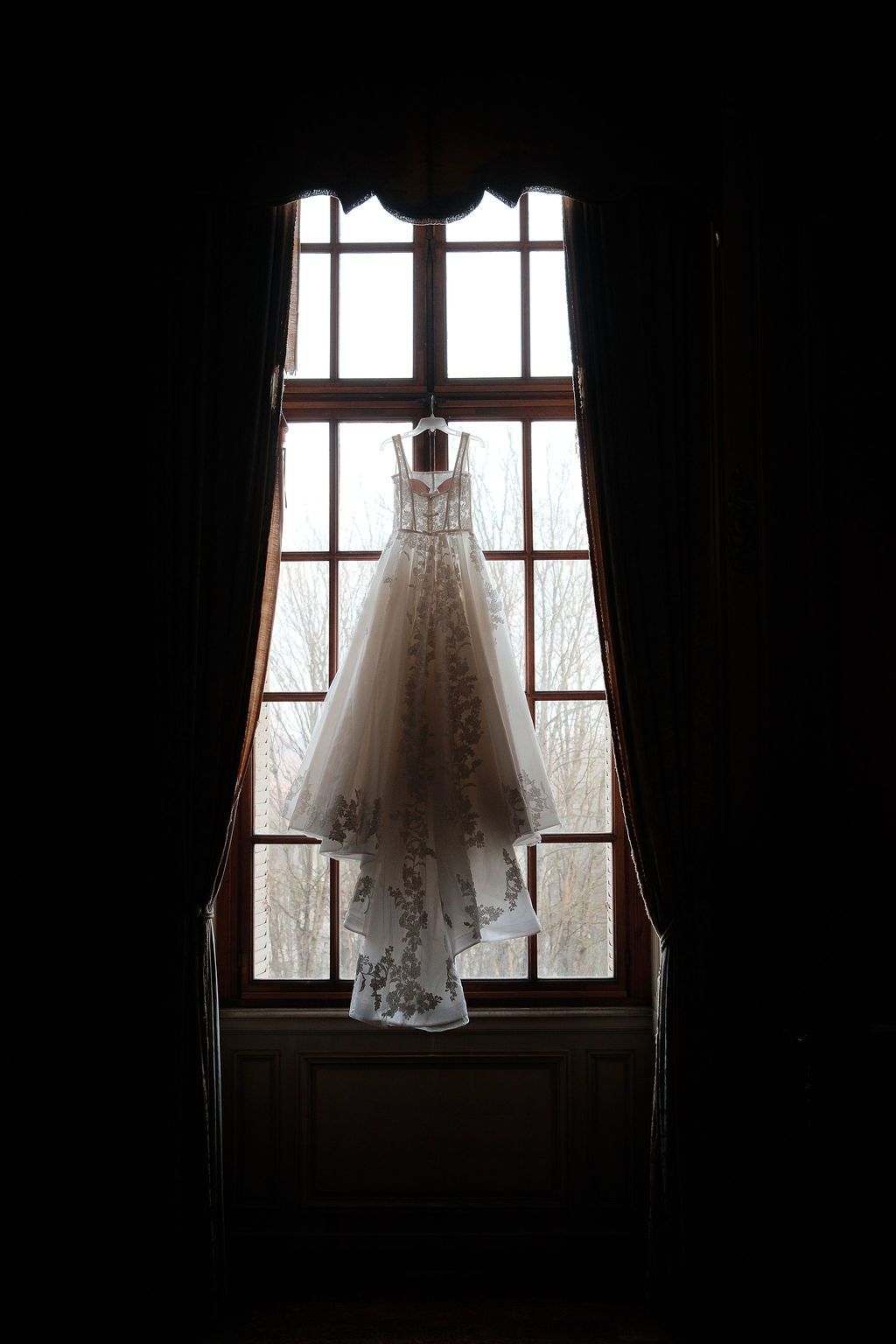 Elegant lace wedding dress hanging in silhouette against tall arched window in dark château room.