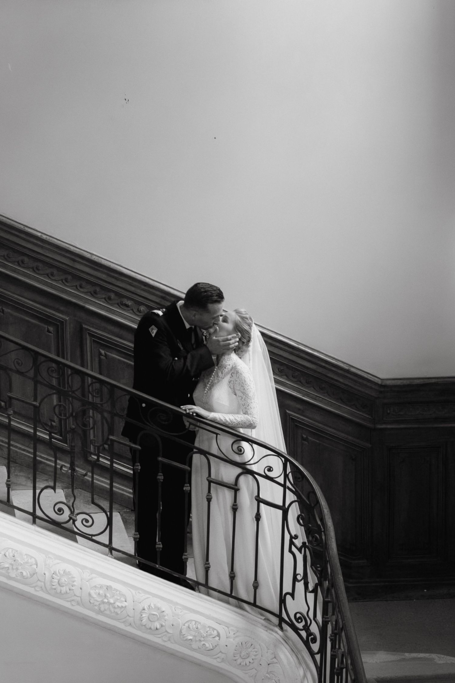 Black and white photo of a bride and groom sharing a kiss on an elegant curved staircase with ornate iron railings.