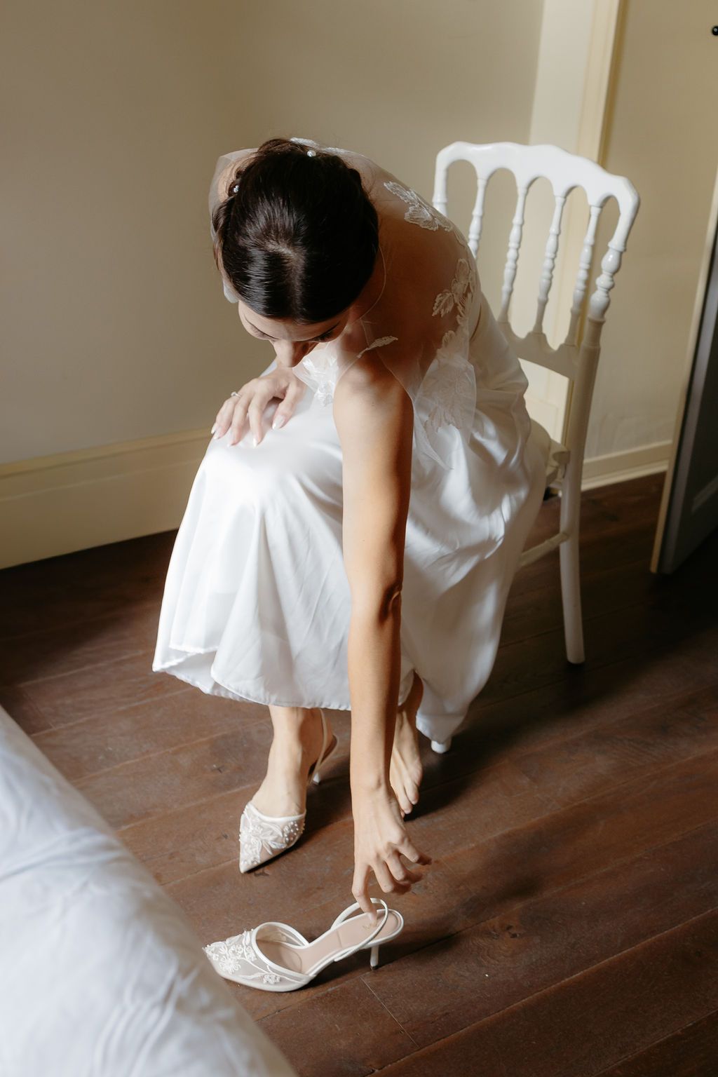 Bride in white lace gown putting on elegant heeled shoes seated on white chair.