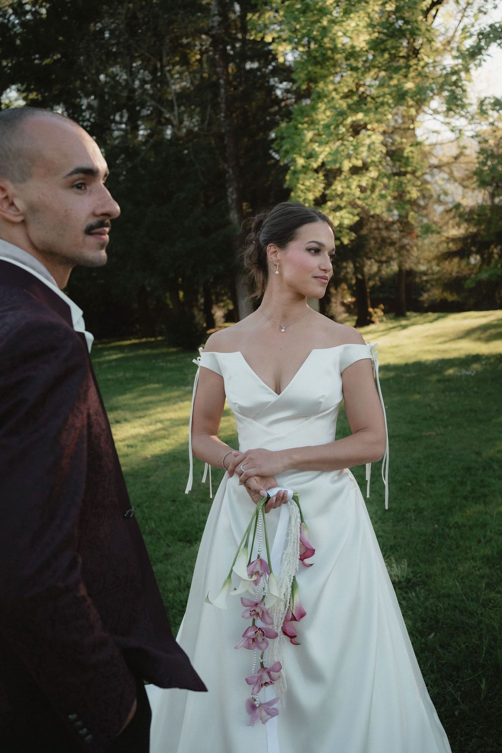 Bride in off-shoulder white gown holds orchid bouquet beside groom in burgundy suit at outdoor wedding.