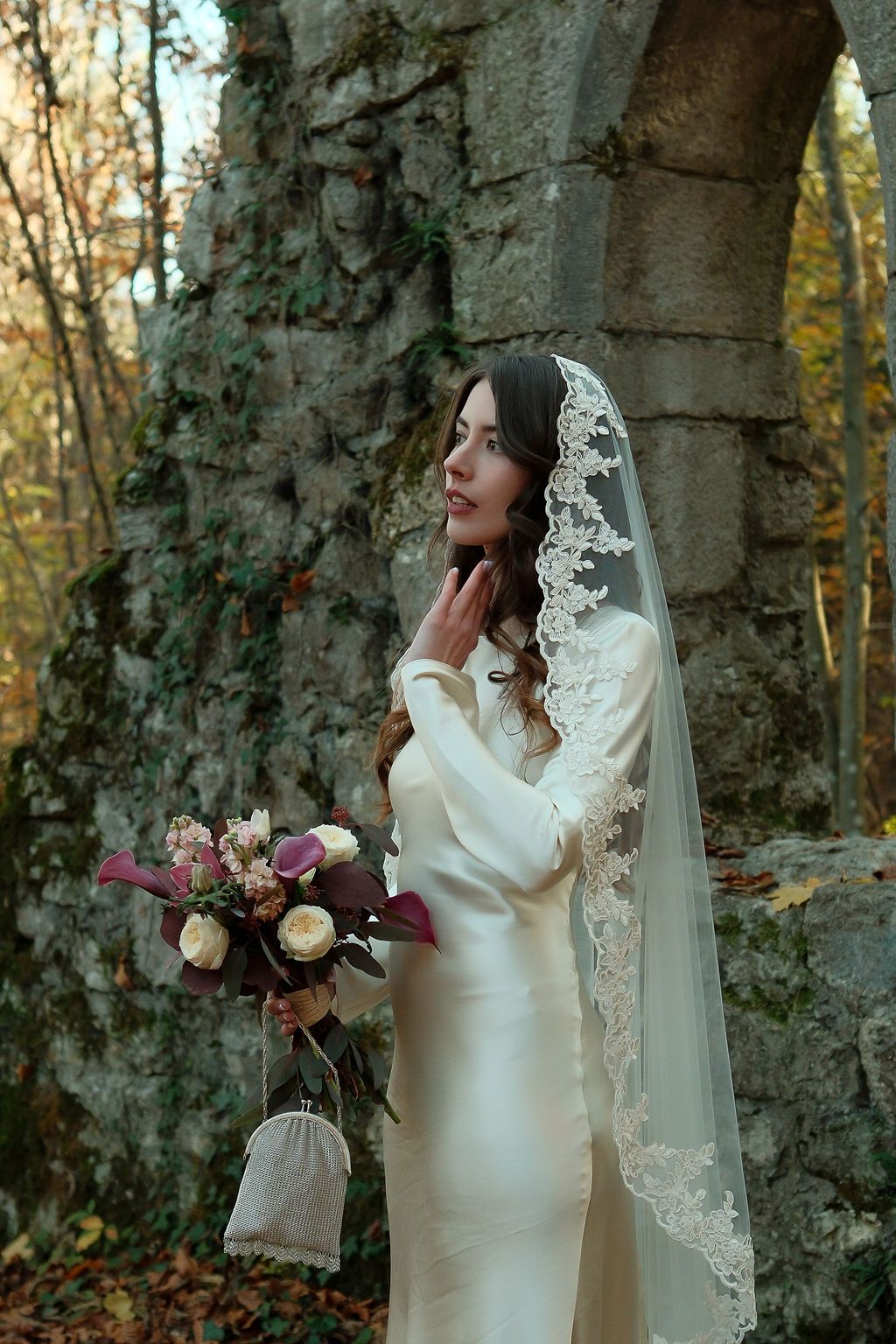 Bride in white gown and lace veil holds bouquet near ivy-covered stone ruins in autumn forest.