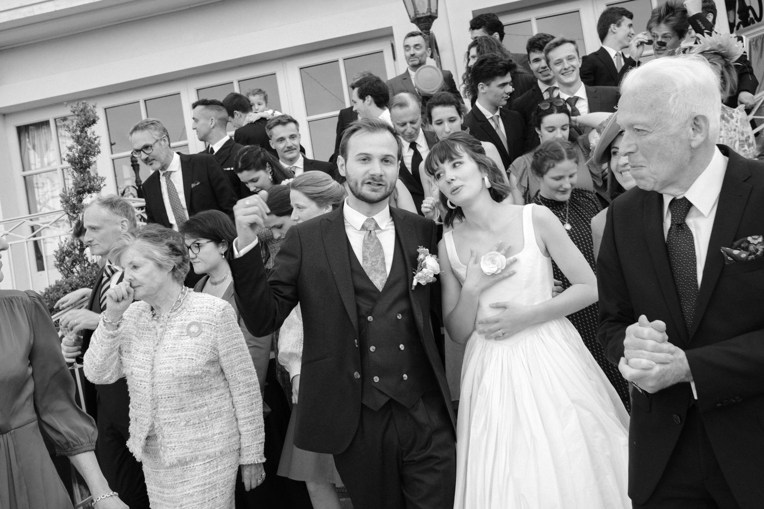 Black and white wedding photo of bride and groom walking with guests celebrating outdoors at a classic venue.