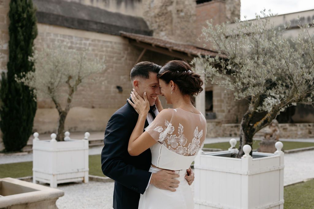 Bride and groom sharing tender embrace in courtyard with olive trees and stone wall backdrop.