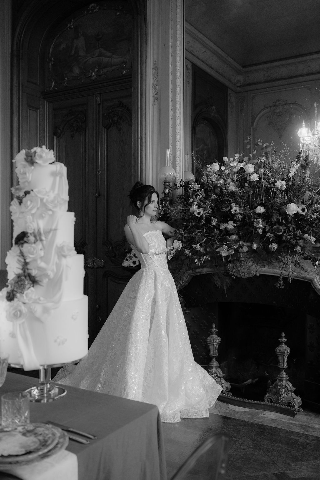 Bride in ballgown standing beside tiered wedding cake near fireplace with floral arrangement.