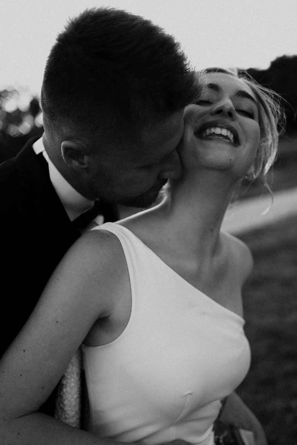 Black and white photo of groom kissing laughing bride on the cheek outdoors.