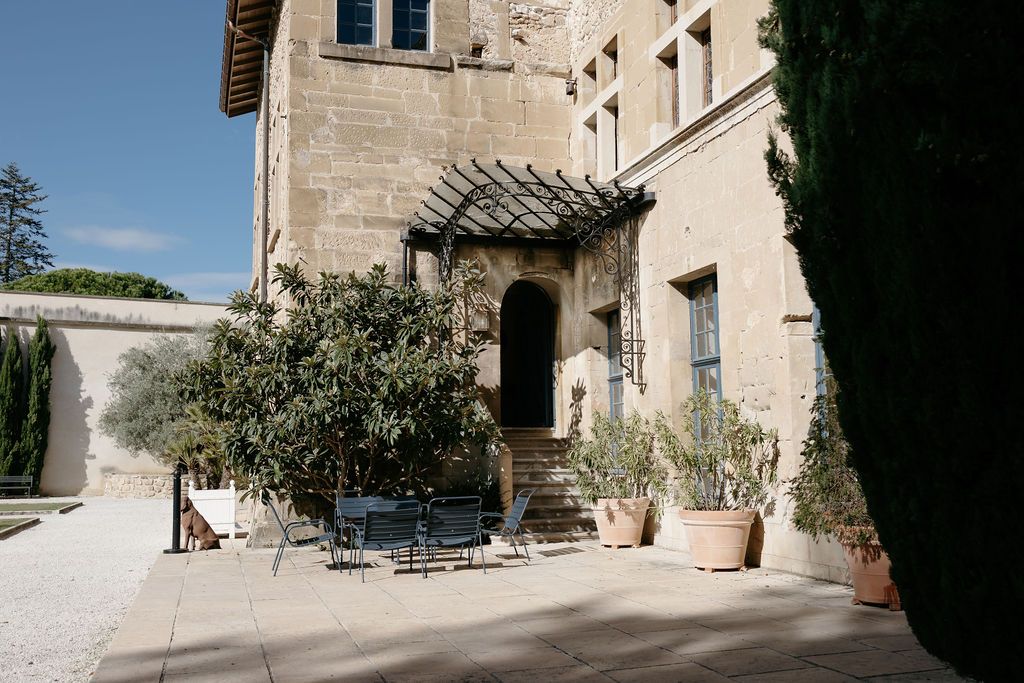 Charming stone French chateau wedding venue exterior with pergola, potted olive trees, and sunny courtyard.