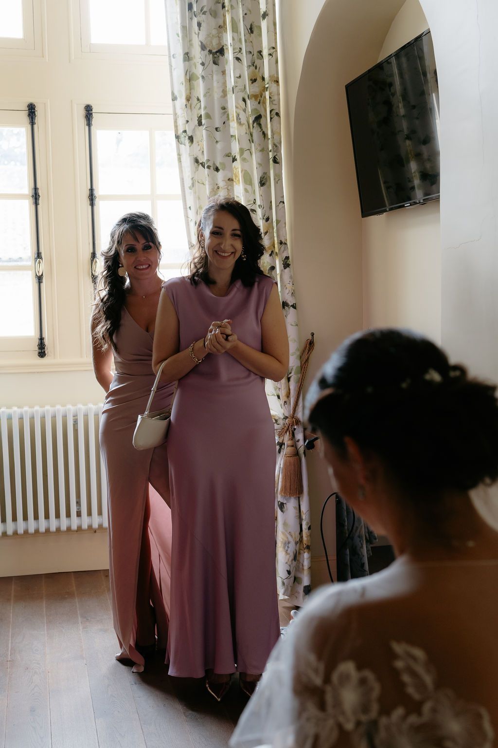 Two bridesmaids in mauve dresses smiling with clasped hands seeing bride for first time.