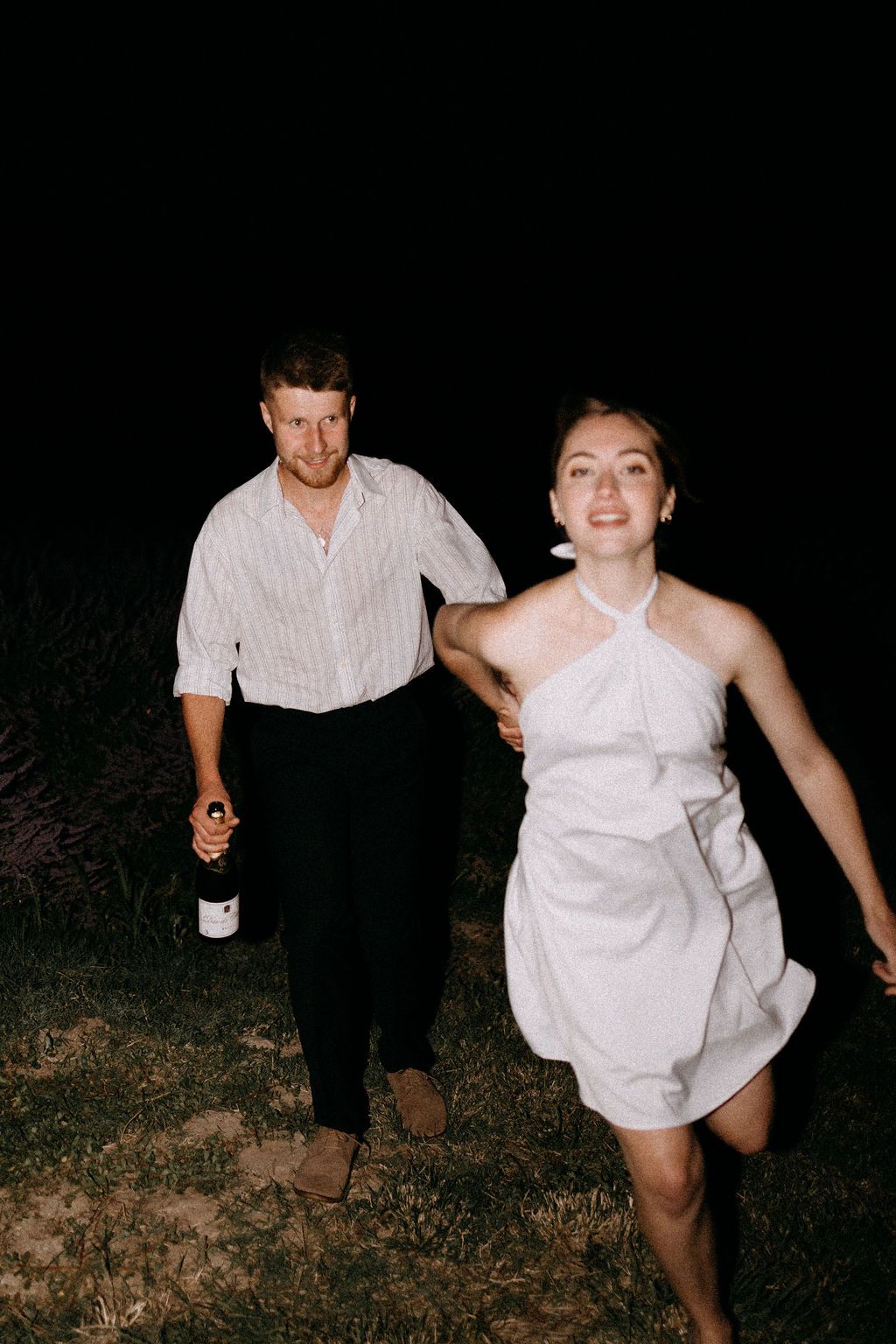 Joyful bride in white halter mini dress runs with groom holding wine bottle through dark outdoor night.
