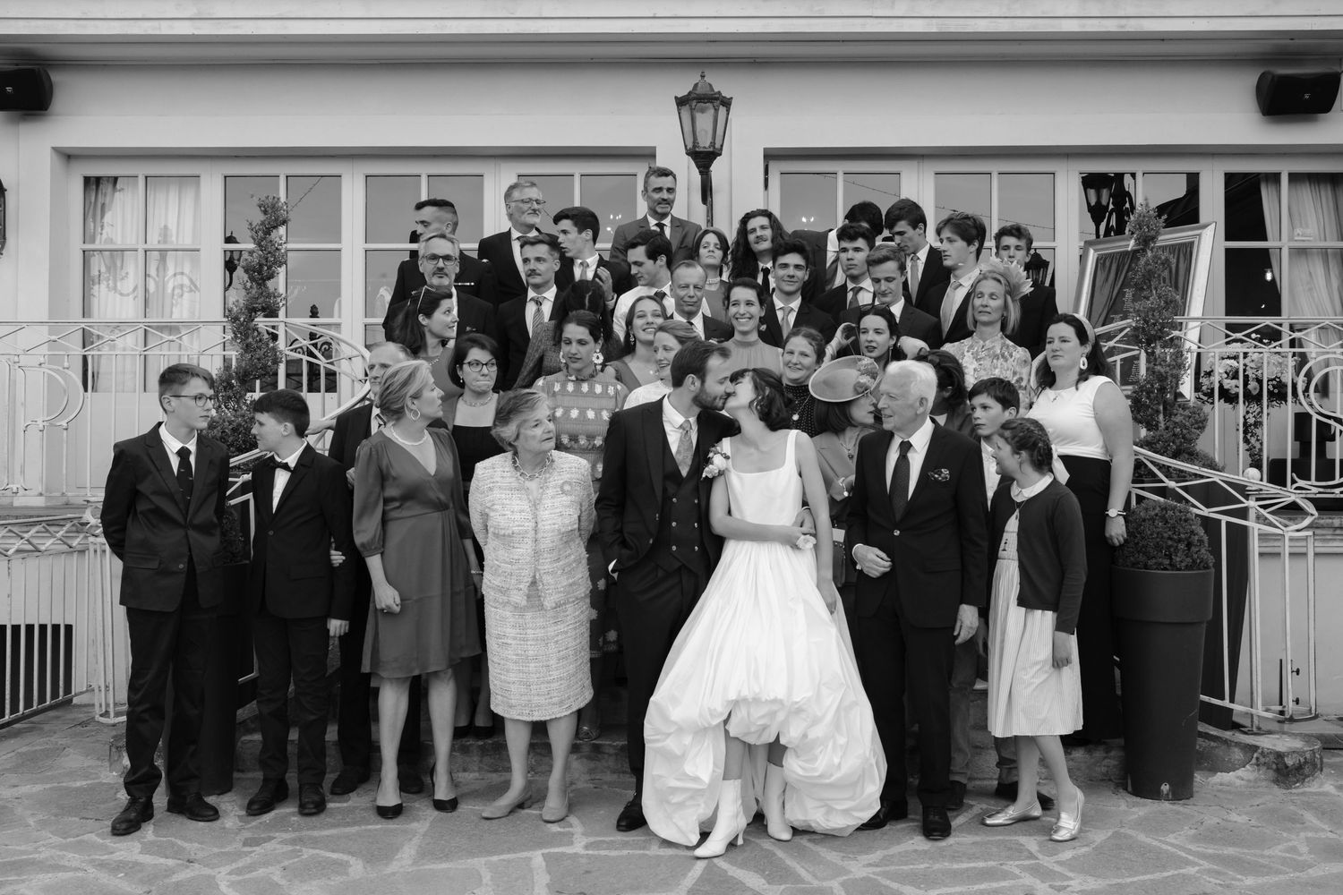 Black and white wedding photo of bride and groom posing with large family group outside elegant venue.