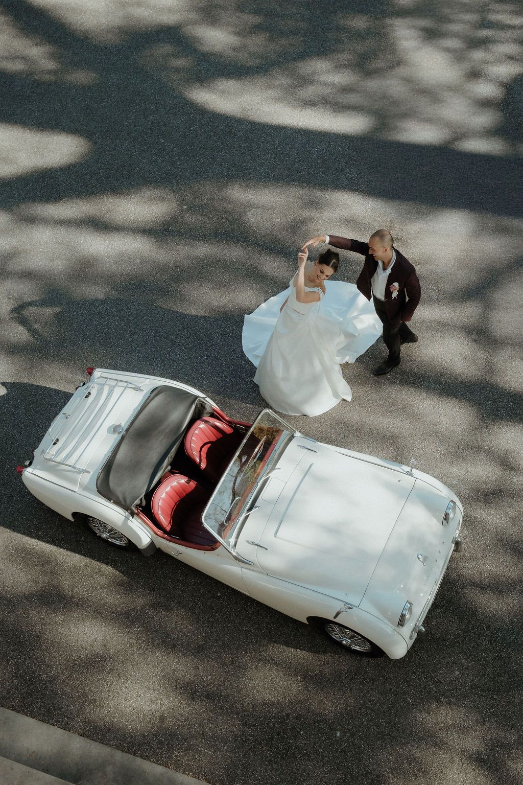 Aerial view of bride and groom dancing beside a vintage white convertible car on a sunny street.