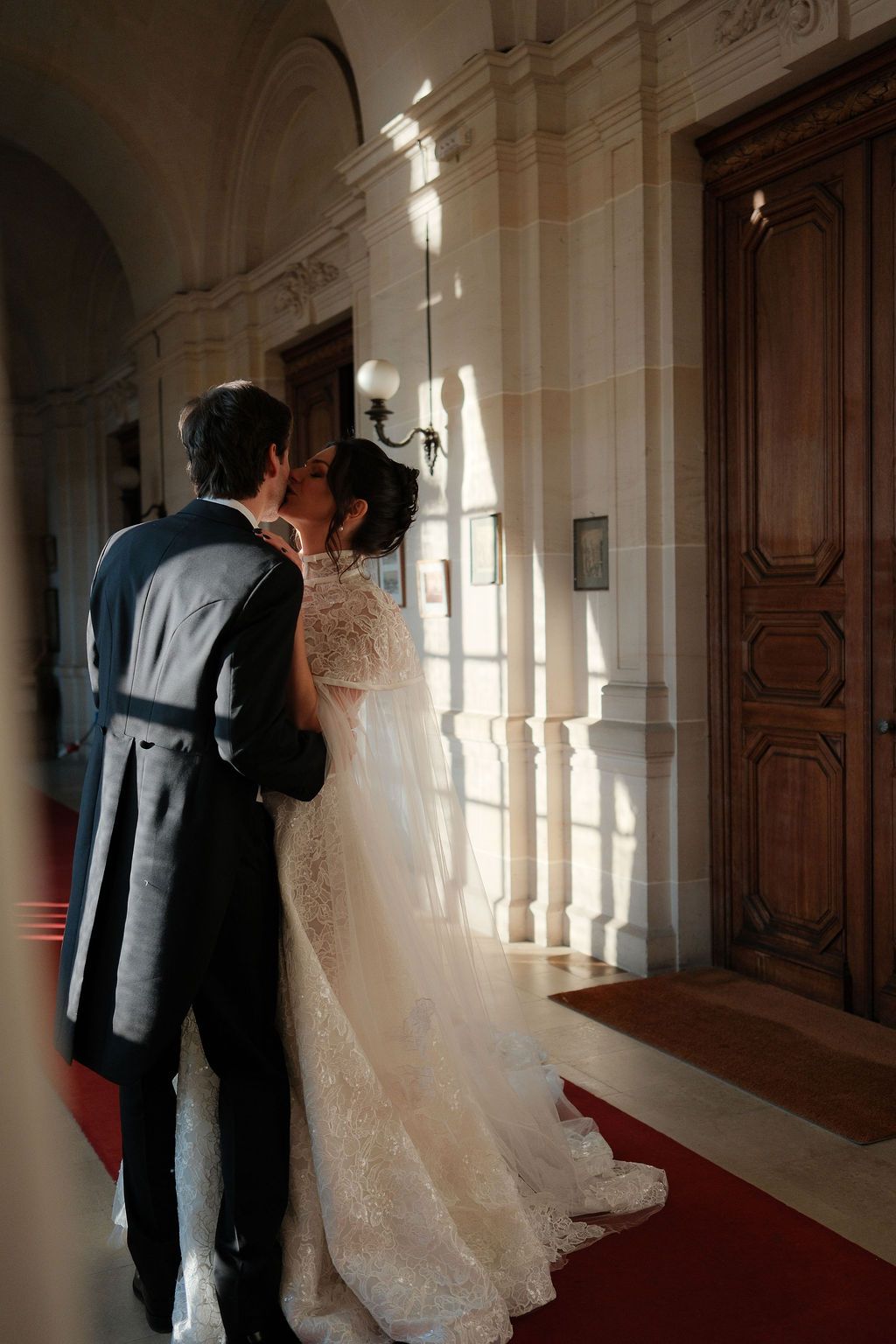 Bride and groom share a romantic kiss in an elegant château corridor with red carpet and ornate stone columns.