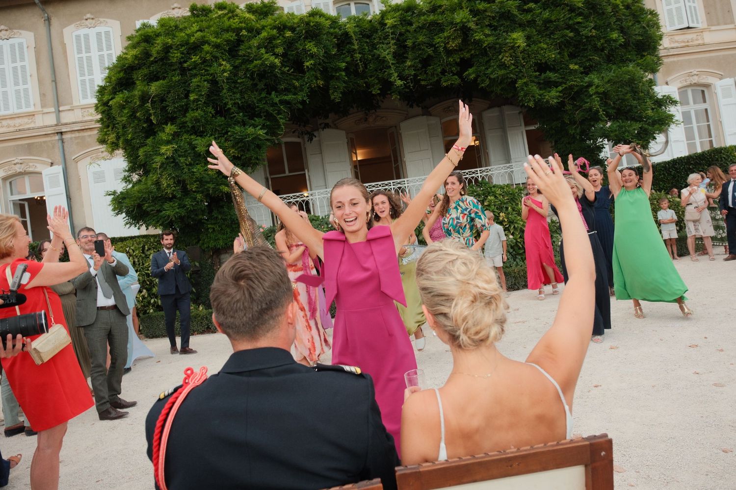 Wedding guests celebrate outdoors, raising arms joyfully near an elegant villa, with women in colorful dresses dancing.