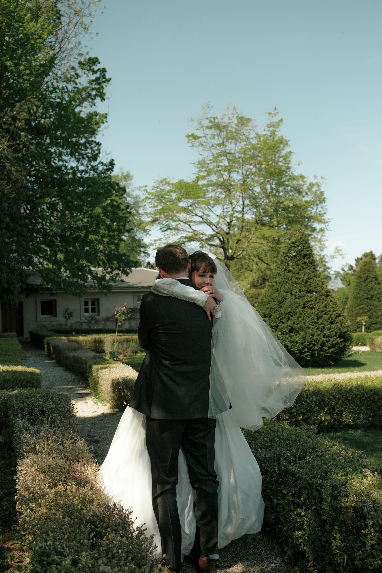 Bride and groom embracing in a lush garden, bride in white gown with veil, groom in dark suit.