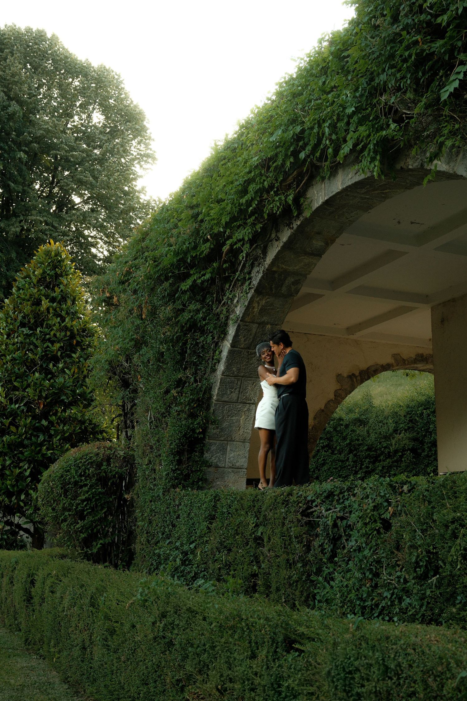 Couple embracing under an ivy-covered stone arch in a lush green garden setting.