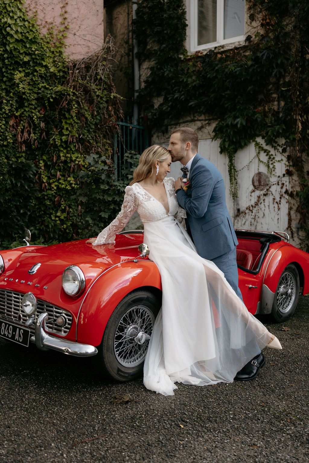 Bride in lace wedding gown sitting on vintage red sports car with groom in blue suit.