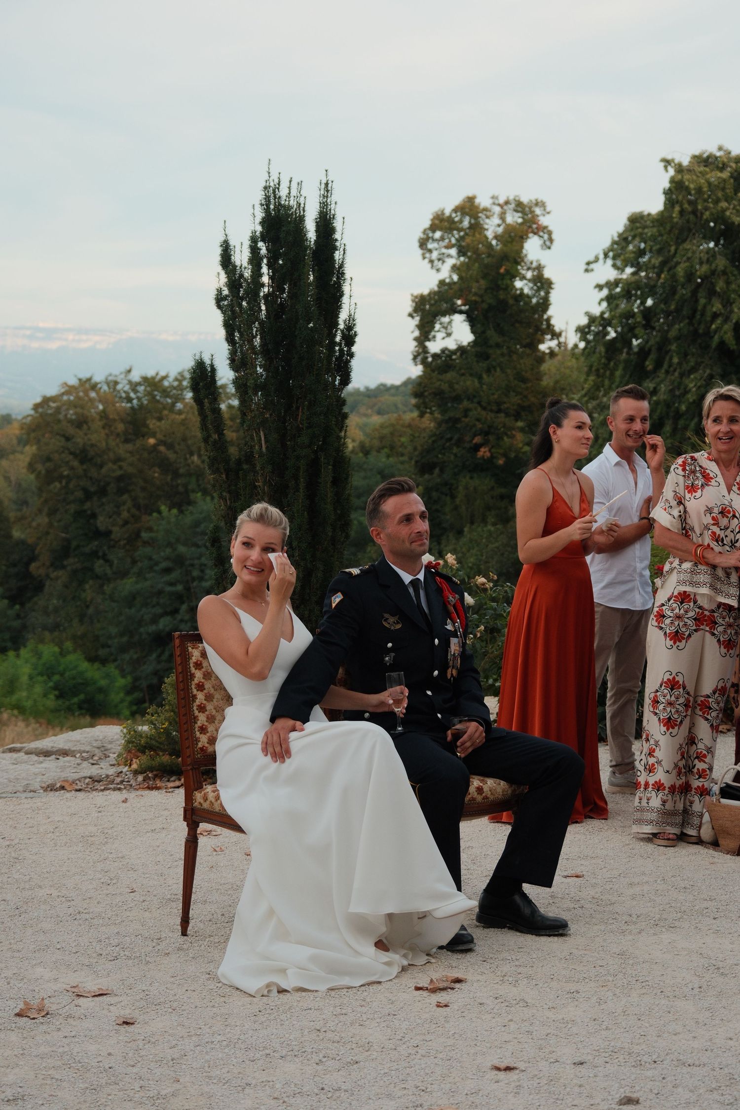 Bride in white dress and groom in military uniform sit together outdoors at wedding ceremony with guests standing behind.