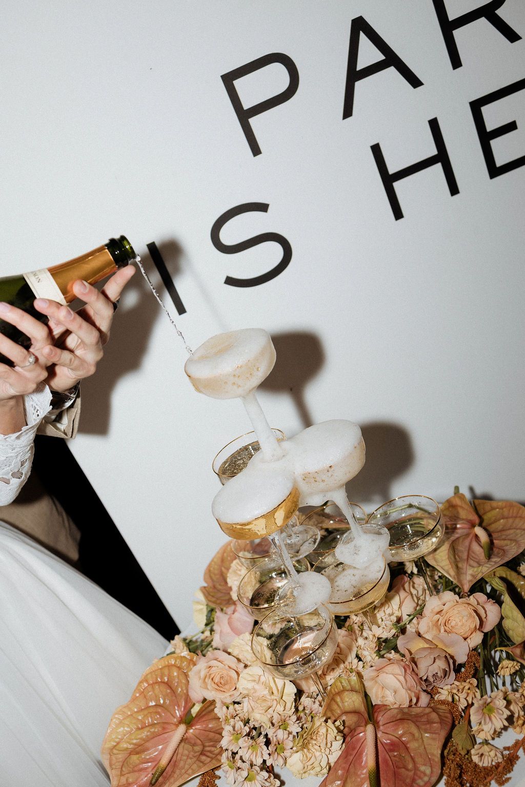 Champagne overflows from wedding glass tower surrounded by pink roses and anthurium flowers at reception.