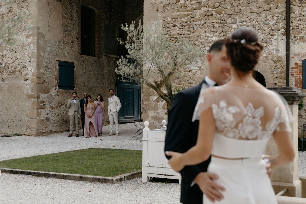 Newlywed couple embracing in courtyard while wedding guests watch near stone wall.