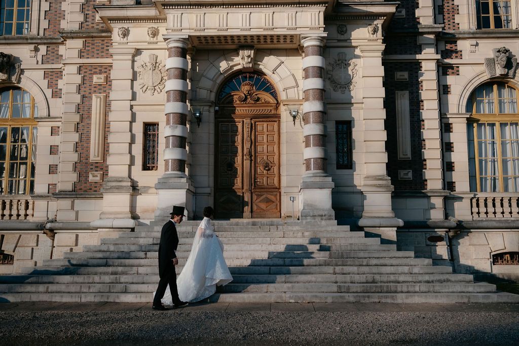 Wedding couple walking toward ornate baroque building entrance in golden sunlight.