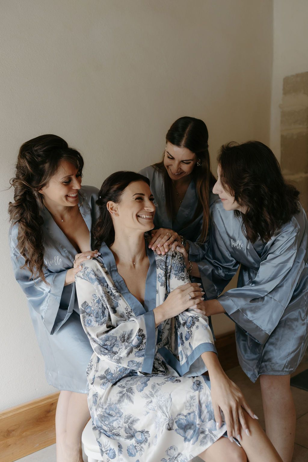 Bridesmaids in blue satin robes laughing with bride in floral robe during wedding morning preparations.