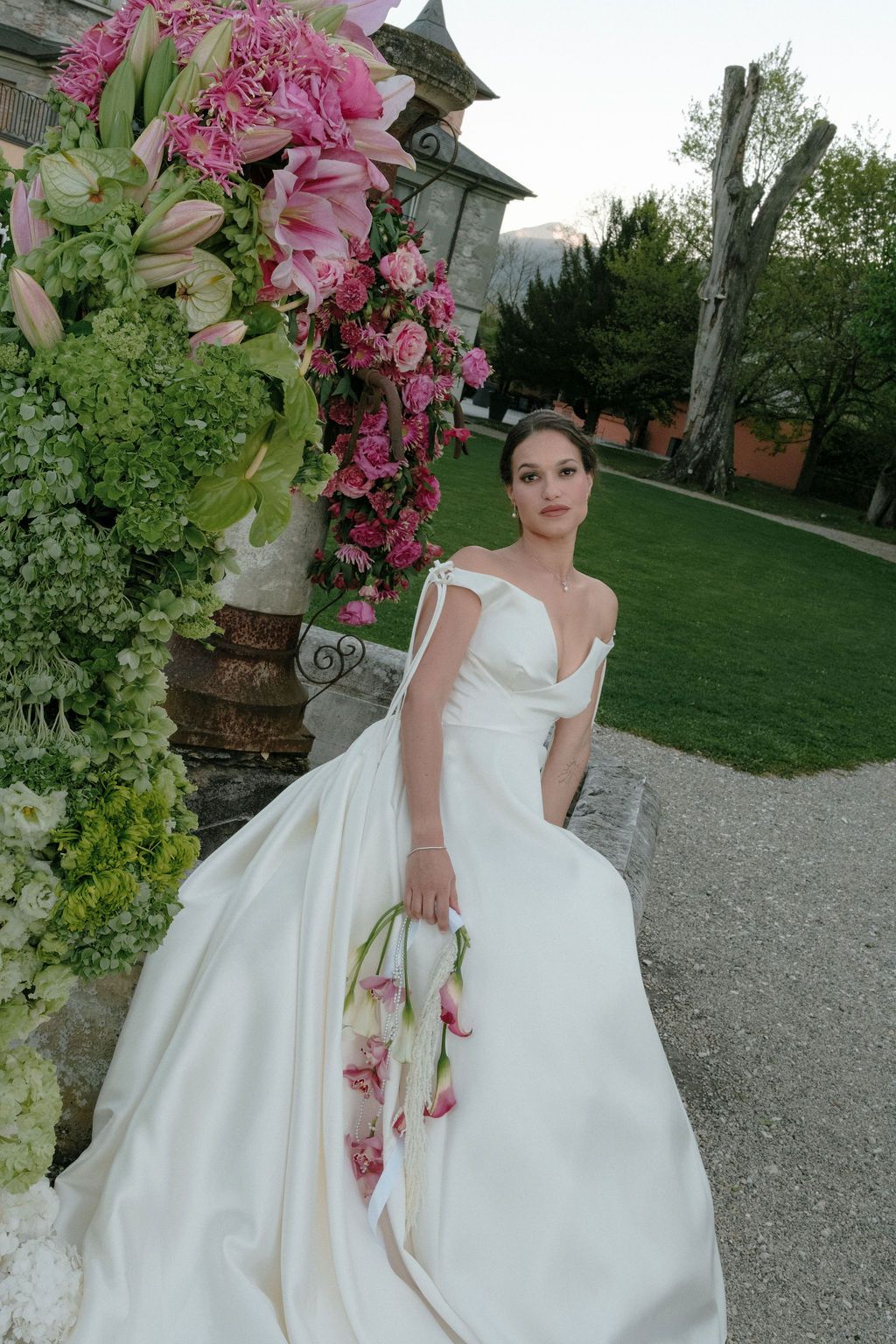 Elegant bride in off-shoulder white gown holding floral bouquet beside lush pink flower arrangement.
