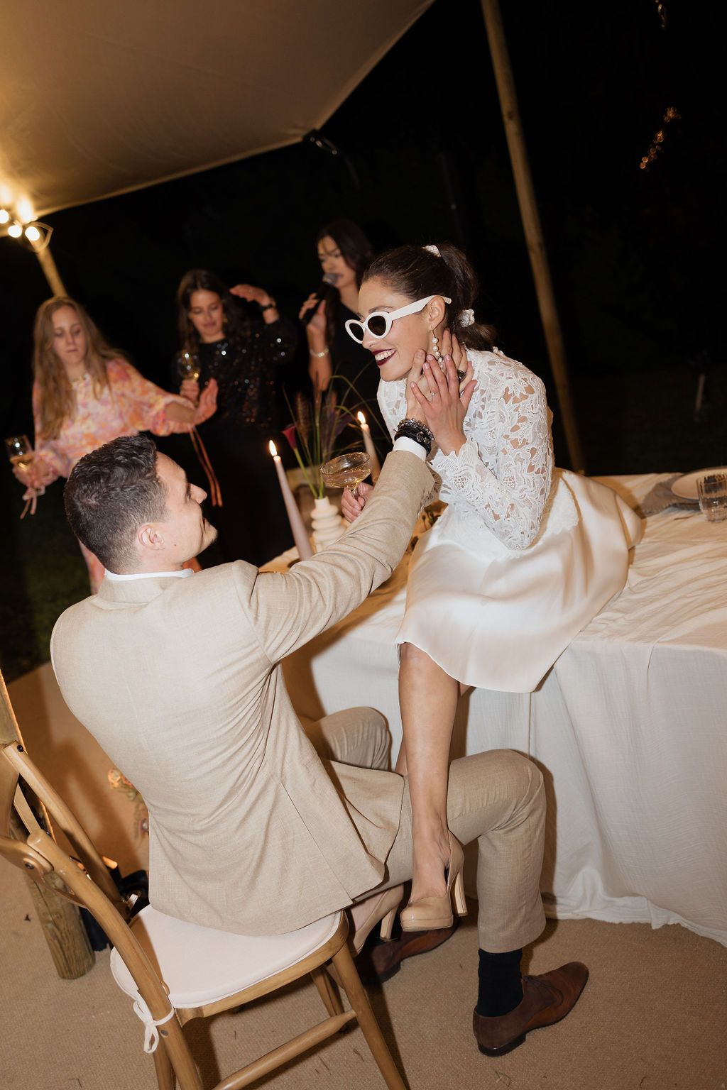 Groom tenderly holds brides face as she laughs in white sunglasses during candlelit outdoor evening reception.