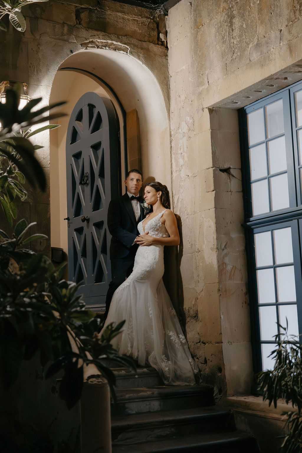 Bride in lace mermaid gown and groom in tuxedo pose in arched doorway of historic stone building.
