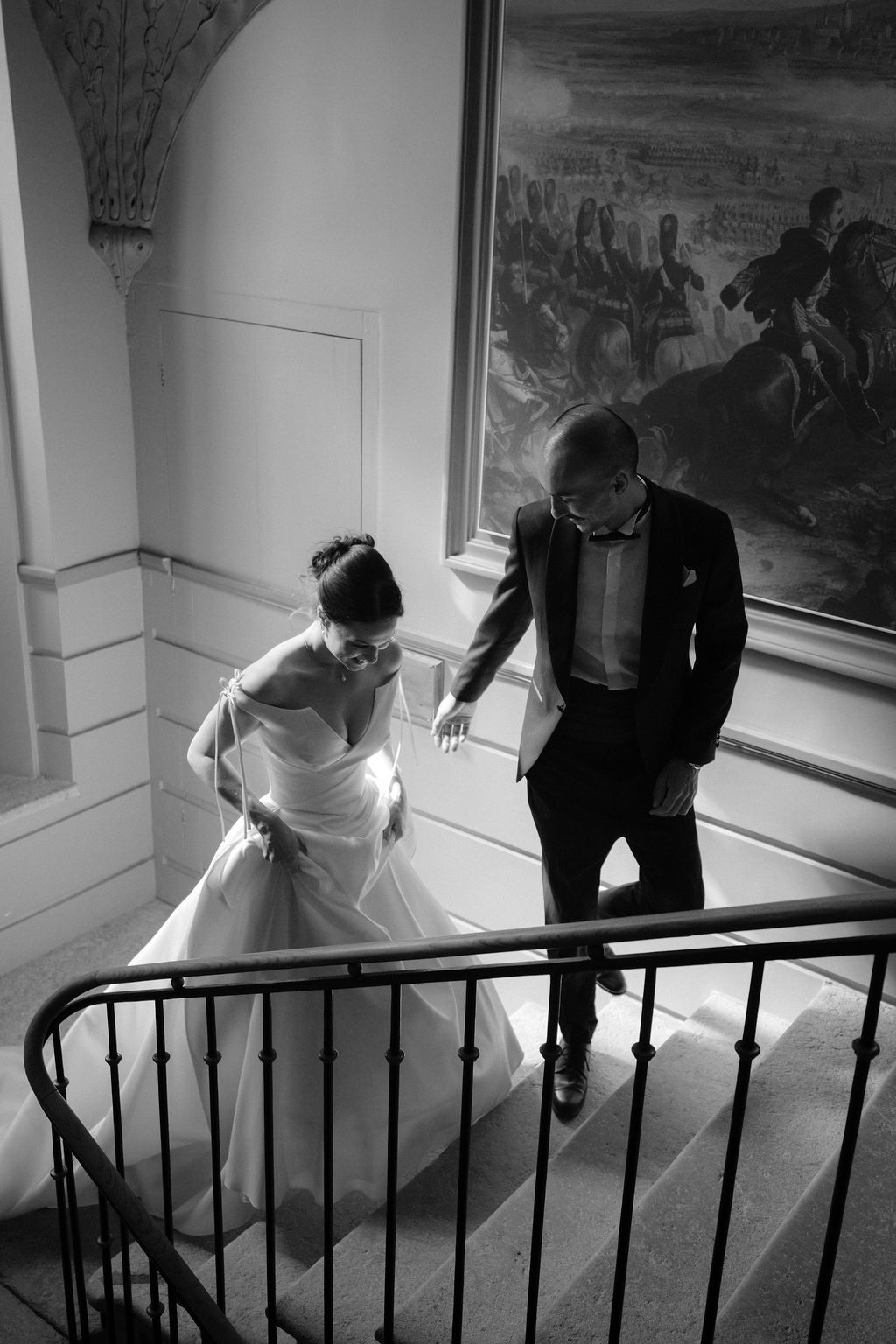 Bride and groom descend elegant staircase in black and white wedding photo with classical painting backdrop.