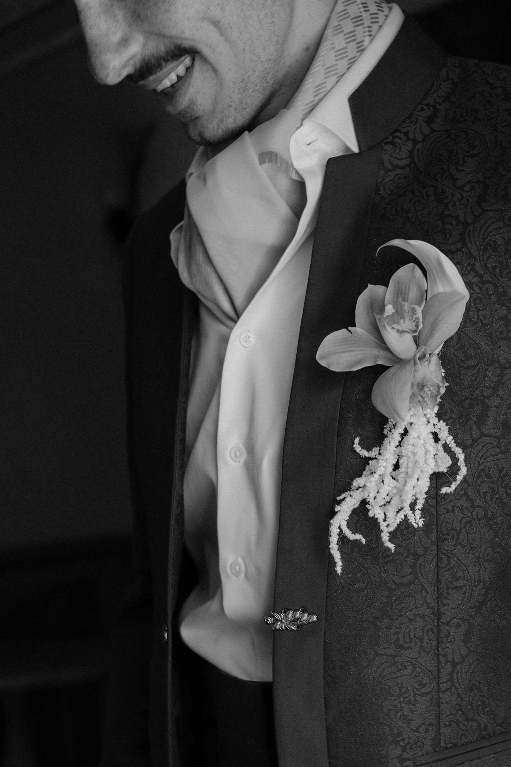 Black and white close-up of groom smiling with orchid boutonniere on embroidered suit jacket lapel.