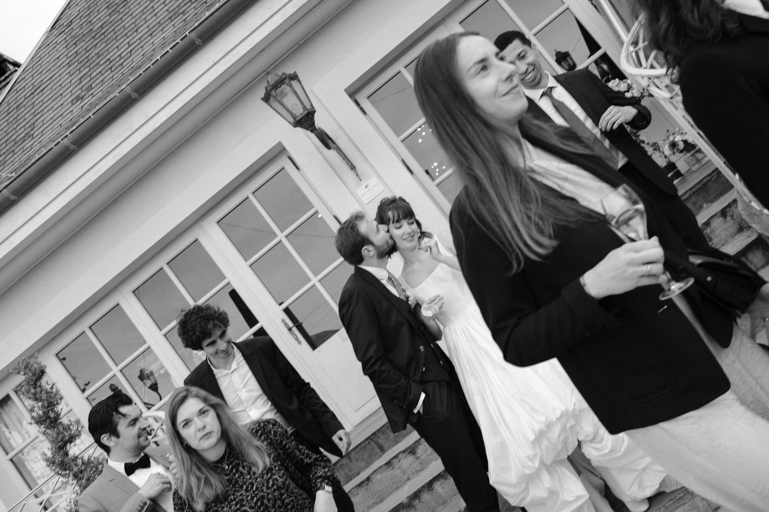 Black and white wedding photo of guests socializing outdoors, with bride and groom kissing in the background.