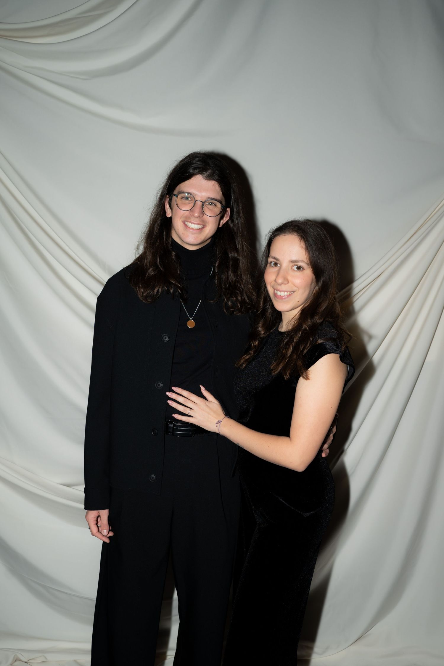 Smiling couple in matching black outfits posing together against a white draped fabric backdrop.