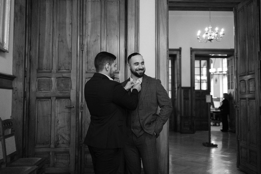 Two groomsmen share a laugh while adjusting boutonnieres in elegant wood-paneled room before wedding ceremony.