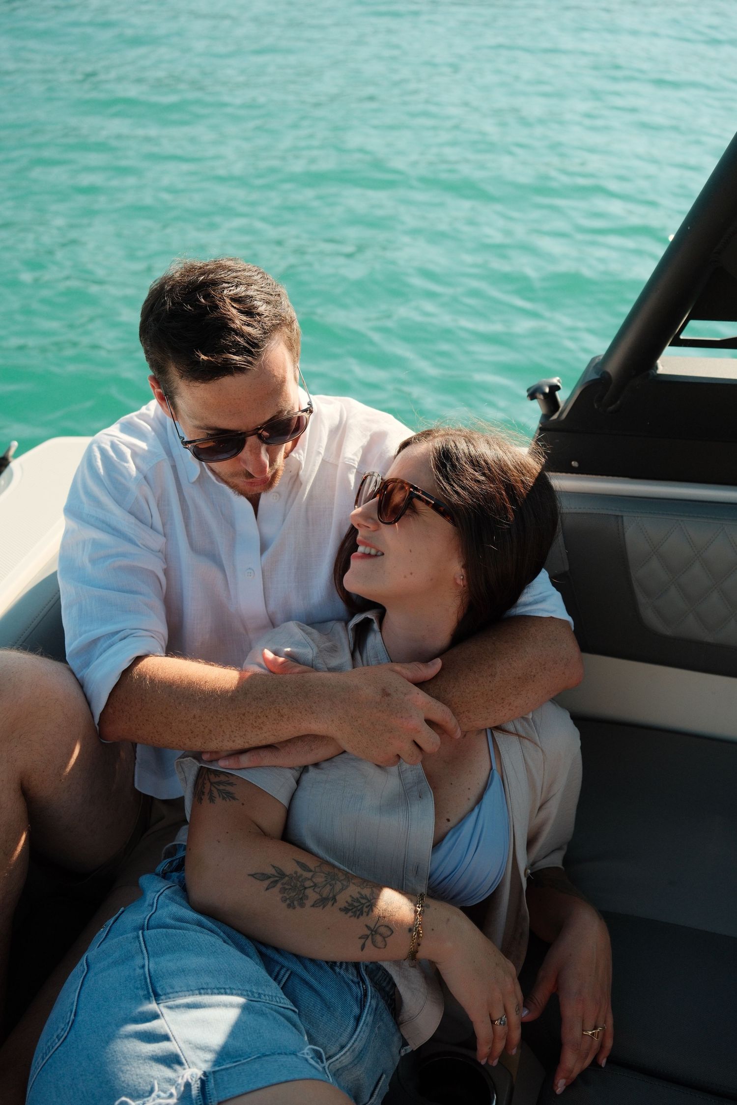 Couple embracing on a luxury boat with turquoise water, wearing sunglasses and casual summer outfits.