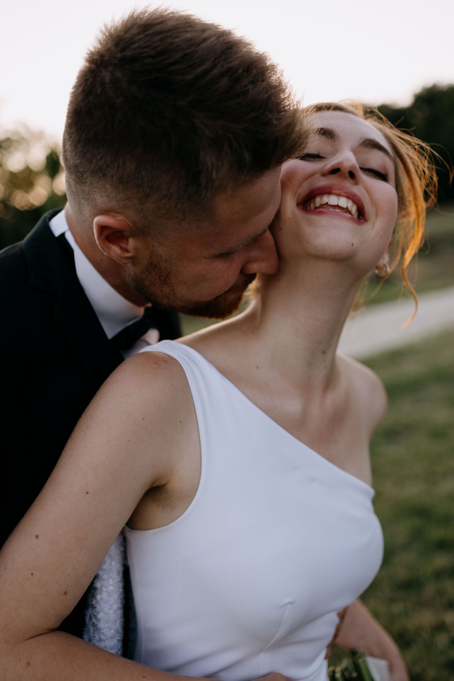 Joyful bride in white dress laughs as groom in black tuxedo kisses her cheek during golden hour.