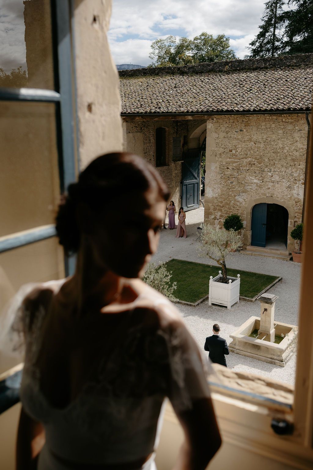 Bride in white wedding dress gazes from window overlooking rustic stone courtyard venue with guests below.