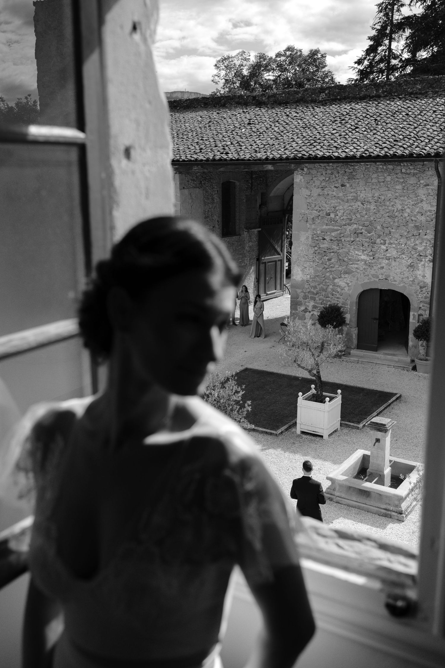 Black and white photo of a bride looking out a window at a courtyard where a groom stands near a fountain.