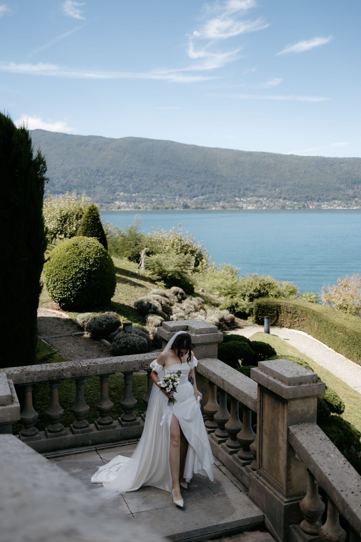 Bride in white gown holding bouquet on stone terrace overlooking scenic lake and mountain landscape on sunny day.