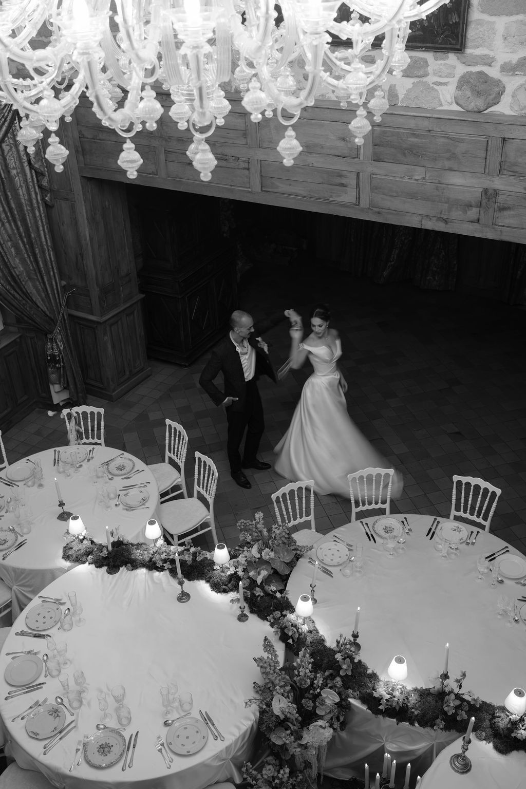 Aerial black and white view of bride and groom dancing in candlelit reception hall with crystal chandeliers.